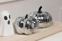 Two decorative pumpkins covered in small, reflective silver mirror tiles, creating a disco ball effect. Each pumpkin has a dark stem and a faceted surface that catches the light. They are displayed on a light wooden tray next to a white ceramic ghost figurine with black eyes and mouth.