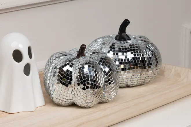 Two decorative pumpkins covered in small, reflective silver mirror tiles, creating a disco ball effect. Each pumpkin has a dark stem and a faceted surface that catches the light. They are displayed on a light wooden tray next to a white ceramic ghost figurine with black eyes and mouth.