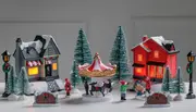 Julelandsbysæt med to oplyste huse, karrusel med rensdyr, miniaturetræer, lygtepæle og figurer. Sne og dekorationer skaber en festlig scene til bordudstilling.