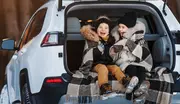 Två barn i vinterjackor och stövlar sitter i en SUV-baklucka, insvepta i en stor rutmönstrad filt, perfekt för utomhusbruk eller resor.