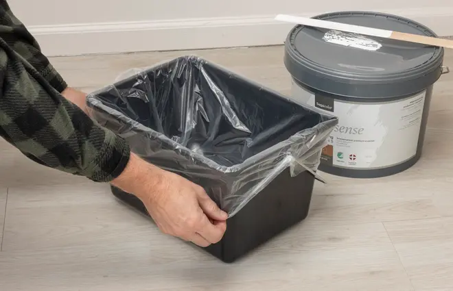 Sort rektangulær affaldsbeholder med monteret plastpose, vist mens den holdes åben af hænder på et lyst gulv.