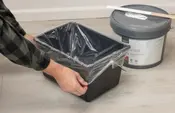 Sort rektangulær affaldsbeholder med monteret plastpose, vist mens den holdes åben af hænder på et lyst gulv.