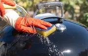 Person med orange handsker rengør sort grill låg med gul svamp og vand, viser grillens indbyggede termometer og robuste metalhåndtag.