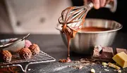 En metalpiskeris dækket af smeltet chokolade over en røreskål, med chokoladebarer, trøfler og bageingredienser på en mørk køkkenoverflade.