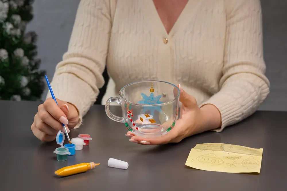 Klar glasmugg med handmålade festliga motiv, inklusive blå stjärna och röda detaljer. Inkluderar små färgburkar, pensel, gul markeringspenna och designskiss på gult papper.
