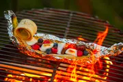Bananbåd dessert i folie, fyldt med skumfiduser, chokolade, blåbær og hindbær, grillet over åben ild. Ideel til lejrbål og udendørs hygge.