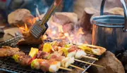 Blandede grillspyd med kød og grøntsager på metalrist over åben ild, hånd med tang og metalgryde ved siden af. Ideel til udendørs madlavning.