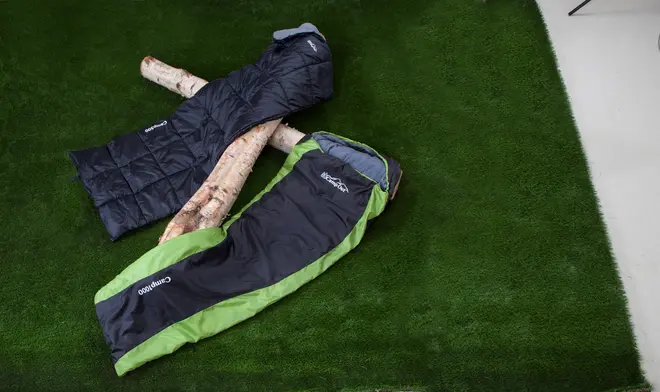 To soveposer i voksenstørrelse, én sort og én sort med grøn kant, vist på kunstgræs. Begge har hætter og er velegnede til camping eller udendørs brug.