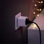 Hvid smart stikprop i en vægkontakt med sort strømledning tilsluttet. Baggrunden viser en mørk væg og lyskæde, hvilket skaber en hyggelig indendørs stemning.