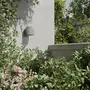 Moderne udendørs væglampe i mat hvid, cylindrisk form, monteret på en enkel væg, omgivet af frodige grønne planter og lyserøde blomster, ideel til have eller terrasse.