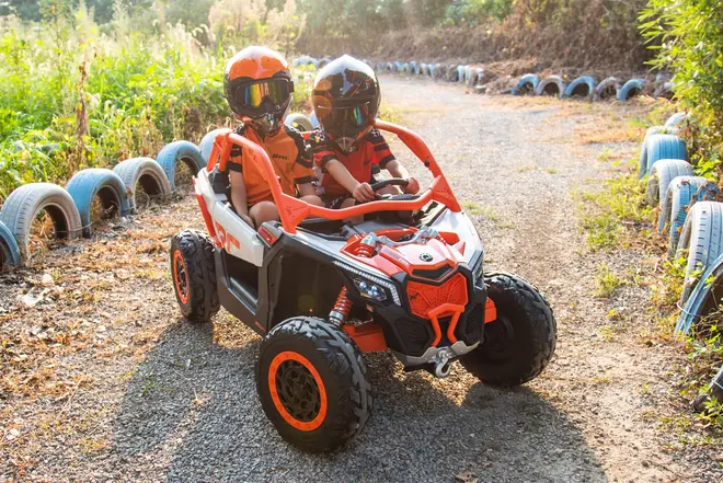 Børns elektrisk køretøj med terrændæk, orange og sort karrosseri, to sæder, rat og sikkerhedssele. Ideel til udendørs leg, realistiske detaljer, robust design, egnet til børn.