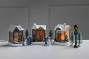 Miniatyr-julby med tre snötäckta hus, träd, lyktstolpe och figurer. Vissa hus är upplysta inifrån och skapar en mysig julstämning.