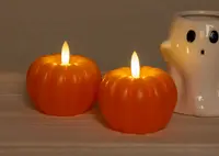 Two orange, pumpkin-shaped candles with realistic flame-shaped LED lights on top, giving a warm, glowing effect. The candles have a smooth, ribbed surface resembling real pumpkins. In the background, there is a white ceramic holder shaped like a ghost with black eyes and mouth, adding a playful, Halloween-themed touch.