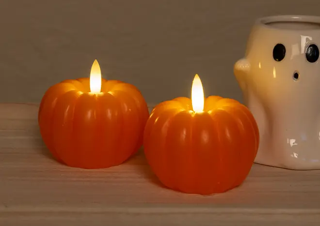 Two orange, pumpkin-shaped candles with realistic flame-shaped LED lights on top, giving a warm, glowing effect. The candles have a smooth, ribbed surface resembling real pumpkins. In the background, there is a white ceramic holder shaped like a ghost with black eyes and mouth, adding a playful, Halloween-themed touch.