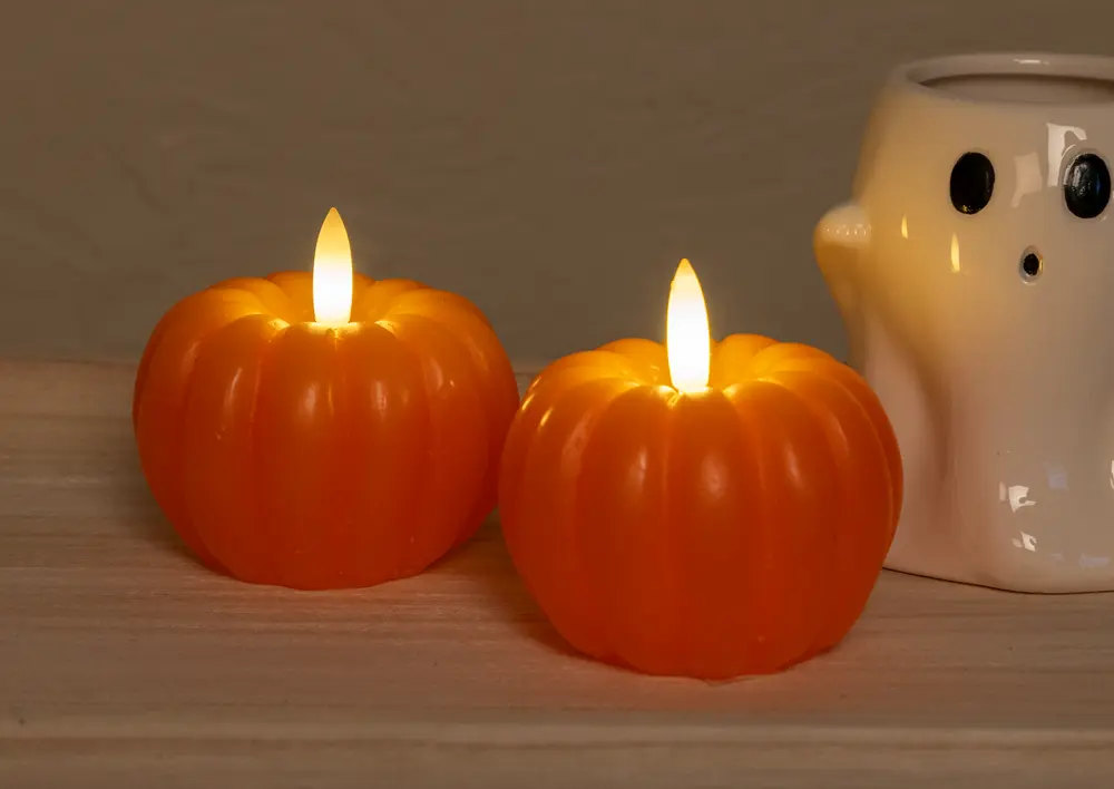 Two orange, pumpkin-shaped candles with realistic flame-shaped LED lights on top, giving a warm, glowing effect. The candles have a smooth, ribbed surface resembling real pumpkins. In the background, there is a white ceramic holder shaped like a ghost with black eyes and mouth, adding a playful, Halloween-themed touch.