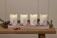 Fyra vita ljus med siffrorna 1 till 4, placerade i en bricka med rosa och silverfärgade kulor, kottar och granris, perfekt för adventsdekoration.