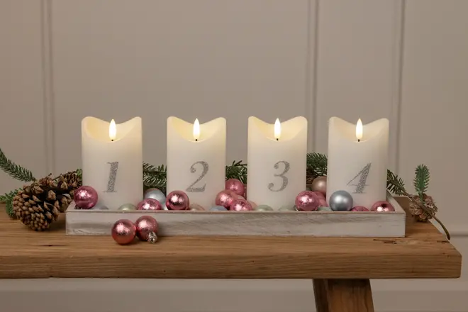 Fyra vita ljus med siffrorna 1 till 4, placerade i en bricka med rosa och silverfärgade kulor, kottar och granris, perfekt för adventsdekoration.