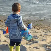 Barn i stribet hættetrøje og gule gummistøvler, holder en blå spand og plastikskovle, står på sandstrand tæt ved vandet. Perfekt til strandleg og udendørs sjov.