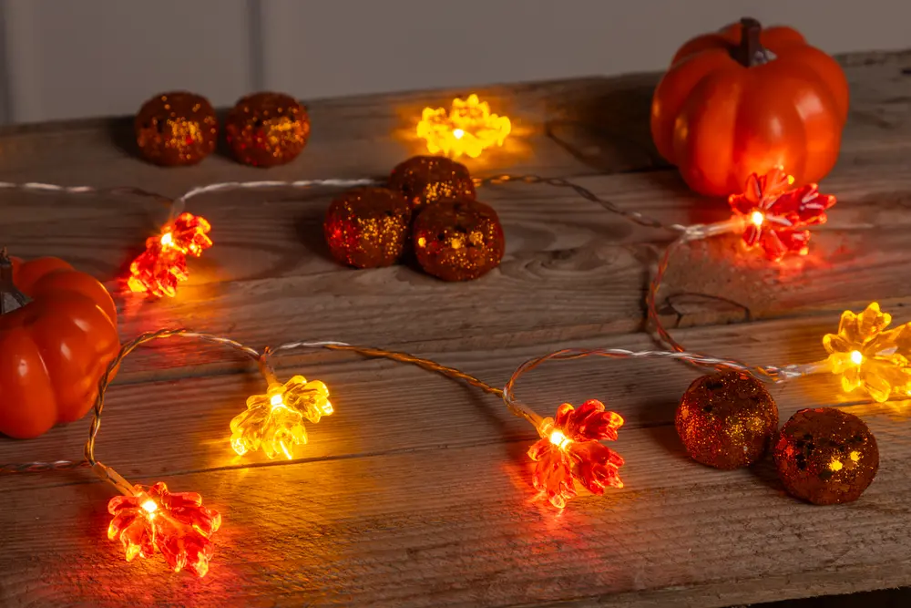 A string of decorative lights featuring translucent maple leaf-shaped covers in warm autumn colors, including orange, red, and yellow. The lights are arranged on a rustic wooden surface, surrounded by small artificial pumpkins and glittery, round ornaments, creating a cozy, festive fall atmosphere.
