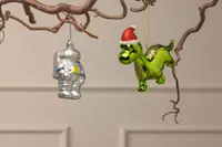Set med två hängande prydnader: en silverfärgad astronaut och en glansig grön dinosaurie med röd tomteluva, båda i glas, perfekta för juldekoration.