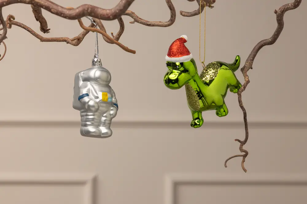 Set med två hängande prydnader: en silverfärgad astronaut och en glansig grön dinosaurie med röd tomteluva, båda i glas, perfekta för juldekoration.