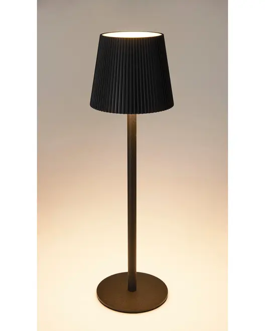 Moderne bordlampe med sort plisseret lampeskærm, slank cylindrisk metalbase og rund flad fod. Udsender varmt lys, velegnet til skrivebord eller natbord.