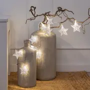 Lyskæde med hvide LED-stjerner med udskårne mønstre, hængt over dekorative keramikvaser og grene. Perfekt til festlig eller hyggelig boligindretning.