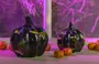 Two glossy black pumpkin-shaped ornaments with a smooth finish, decorated with shiny gold bat motifs. The pumpkins have a realistic ribbed texture and short, thick stems. Surrounding them are small, glittery orange balls, adding a festive touch. The background features a window with purple lighting, creating a vibrant and spooky atmosphere.