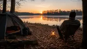 Lejrscene ved sø med kuppeltelt, lanterne, klapstol og træstub, mens solen står op over det stille vand. - Billedet er skabt med AI