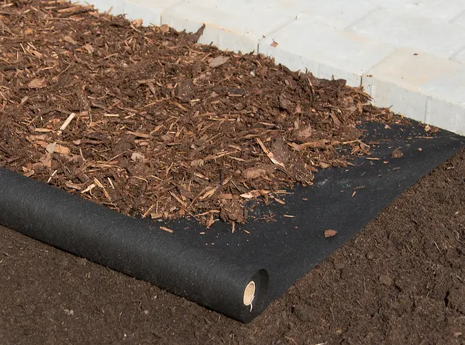 Svart marktäckningsduk utrullad på jord, används som ogräsbarriär under täckbark i rabatter. Synlig täckbark och marksten i bakgrunden.