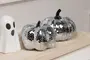 Two decorative pumpkins covered in small, reflective silver mirror tiles, creating a disco ball effect. The pumpkins have dark stems and a glossy, faceted surface. They are displayed on a light wooden tray next to a small white ceramic ghost with black eyes and mouth.