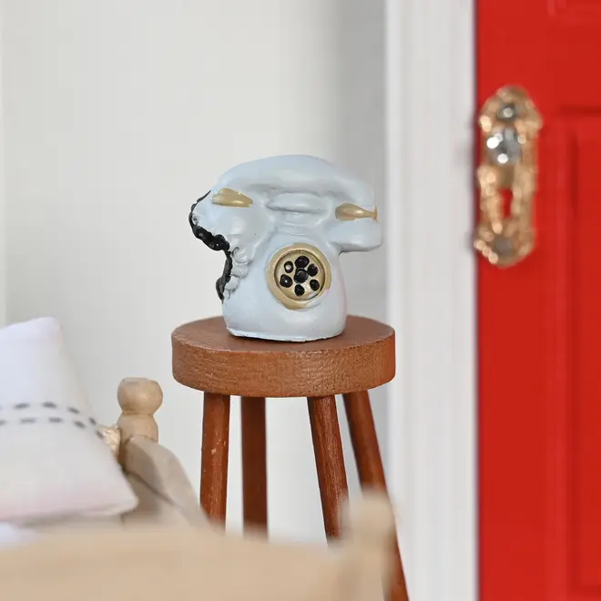 Miniature retro drejeskivetelefon, lyseblå med guld- og sort detaljer, placeret på en træskammel. Perfekt til dukkehusindretning eller samlerdisplay. Realistisk, ikke-funktionel.