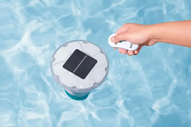 Flydende solcelledrevet poollys med sort solpanel på toppen, vist i en pool. En hånd holder en fjernbetjening til at styre lyset.