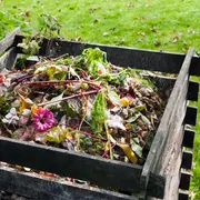Udendørs kompostbeholder i træ fyldt med organisk affald som grøntsagsrester, frugtskræller, visne blomster og haveaffald, velegnet til hjemmekompostering og bæredygtig havebrug.