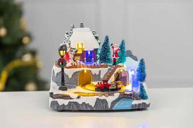 Oplyst julelandsbydekoration med snedækket hus, julemand, grantræer, tog på skinner, gadelygte og lille vandfald. Festlig og detaljeret julescene.