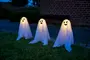 Set med tre utomhus Halloween-spökdekorationer med lysande lampor, vita tygstycken och svarta ansikten. Varje spöke har olika ansiktsuttryck och står på gräs.
