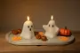 Two white candles shaped like cartoon ghosts, each with black eyes and a mouth, are lit and placed on a light-colored stone tray. The tray is decorated with small orange glittery balls, flat pumpkin-shaped cutouts, and a small artificial pumpkin, creating a festive Halloween-themed display.
