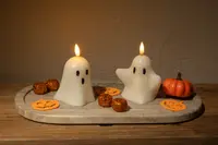 Two white candles shaped like cartoon ghosts, each with black eyes and a mouth, are lit and placed on a light-colored stone tray. The tray is decorated with small orange glittery balls, flat pumpkin-shaped cutouts, and a small artificial pumpkin, creating a festive Halloween-themed display.