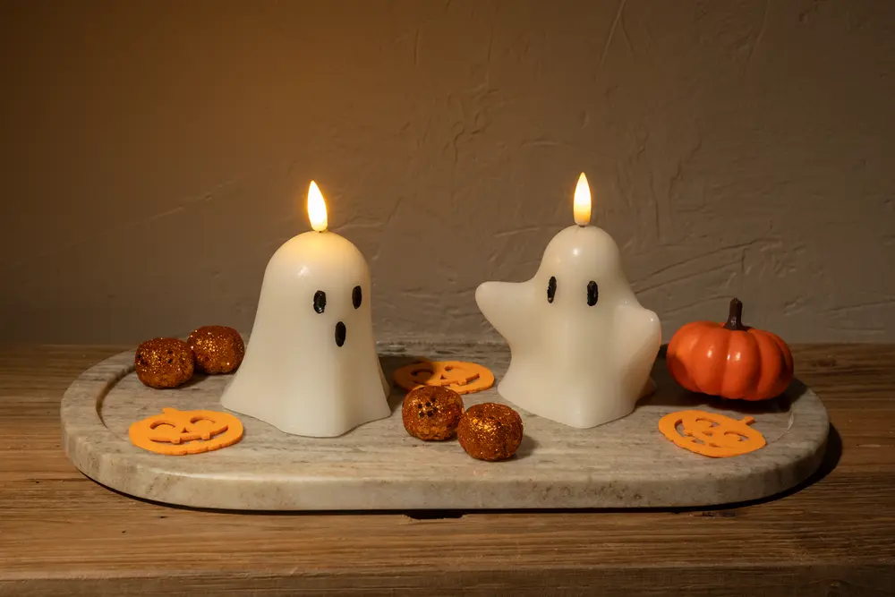 Two white candles shaped like cartoon ghosts, each with black eyes and a mouth, are lit and placed on a light-colored stone tray. The tray is decorated with small orange glittery balls, flat pumpkin-shaped cutouts, and a small artificial pumpkin, creating a festive Halloween-themed display.