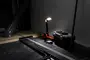 Bærbar LED arbejdslampe med justerbart hoved, rød base og sort håndtag. Placeret ved siden af sort værktøjskasse, perfekt til arbejdsområder eller byggepladser.