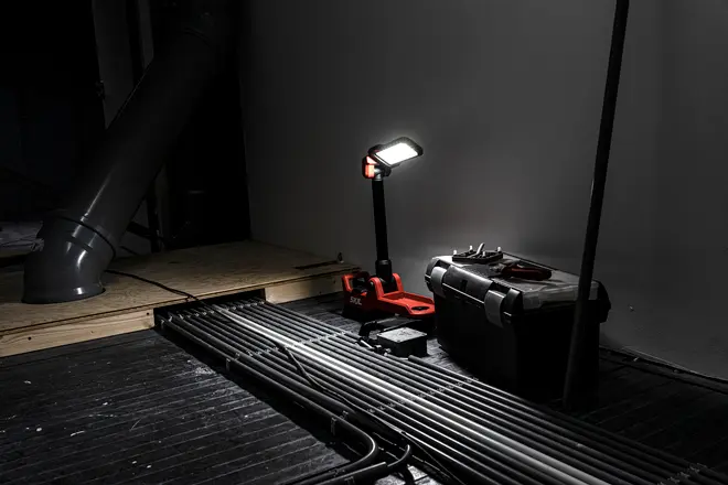 Bærbar LED arbejdslampe med justerbart hoved, rød base og sort håndtag. Placeret ved siden af sort værktøjskasse, perfekt til arbejdsområder eller byggepladser.