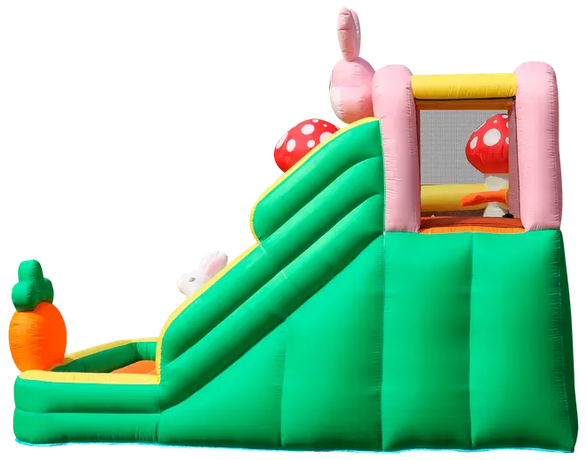 Oppustelig grøn og gul rutsjebane med lyserøde kanin- og svampedekorationer, gulerodsformet oppustelig ved bunden og netindhegning øverst. Til børns udendørs leg.