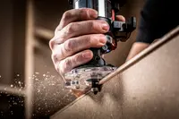 Handhållen elektrisk träfräs med genomskinlig bas, används för att forma och trimma träkanter. Ergonomiskt grepp, synlig fräs och träspån under arbete.