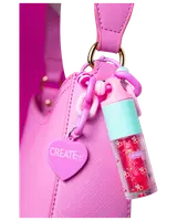 Lyserød håndtaske med hjerteformet "CREATE it!" vedhæng og farverig kæde, der holder en lille lipgloss med turkis låg og kirsebærmønster.