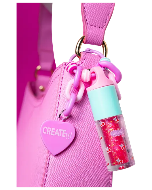 Lyserød håndtaske med hjerteformet "CREATE it!" vedhæng og farverig kæde, der holder en lille lipgloss med turkis låg og kirsebærmønster.