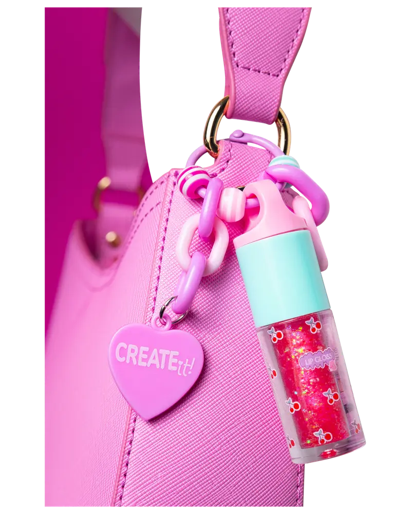Lyserød håndtaske med hjerteformet "CREATE it!" vedhæng og farverig kæde, der holder en lille lipgloss med turkis låg og kirsebærmønster.