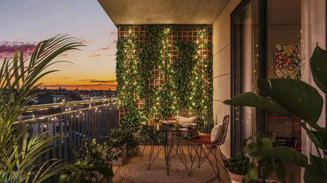 Hyggelig altan med rundt bord, to stole, grønne planter, lyskæder og lodret havevæg. Solnedgangsudsigt, moderne indretning, ideel til udendørs spisning eller afslapning.