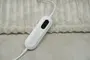Elektrisk varmetæppe-controller med tre knapper og digitalt display, fastgjort til et blødt, lysebeige tæppe. Bruges til at justere varmeindstillinger og tænd/sluk-funktioner.