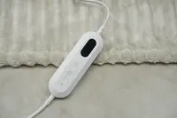 Elektrisk värmefiltkontroll med tre knappar och digital display, fäst vid en mjuk, ljusbeige filt. Används för att justera värme och slå på/av.