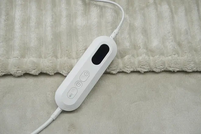 Elektrisk varmetæppe-controller med tre knapper og digitalt display, fastgjort til et blødt, lysebeige tæppe. Bruges til at justere varmeindstillinger og tænd/sluk-funktioner.